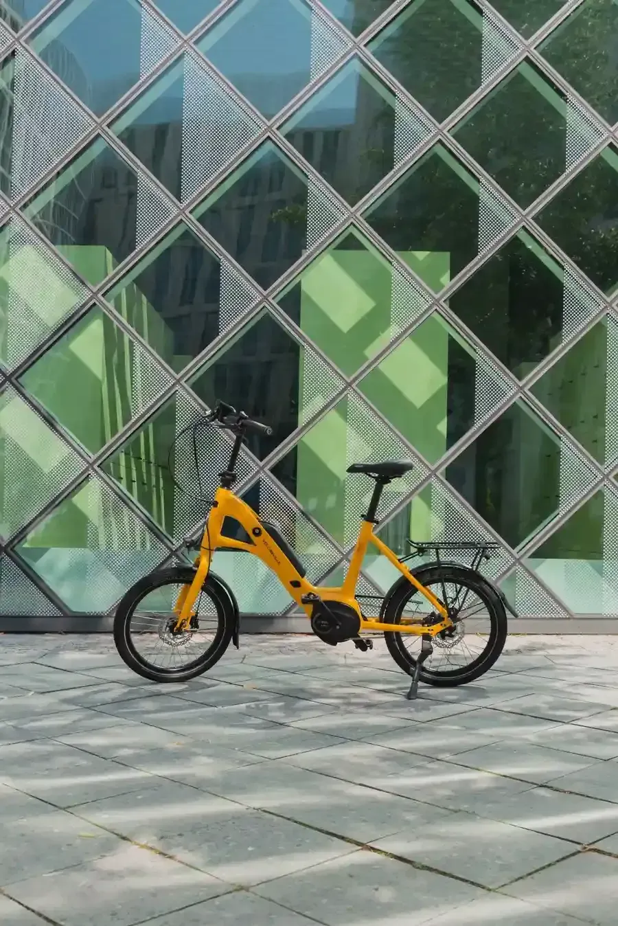Bicicleta naranja aparcada en una acera de baldosas frente a una pared de cristal con estampado de rombos verdes.