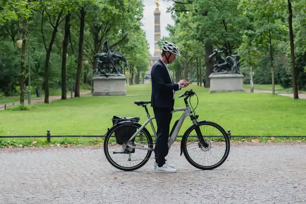 Una persona de pie con una bicicleta en un parque, mirando un teléfono, con árboles verdes y un monumento al fondo.