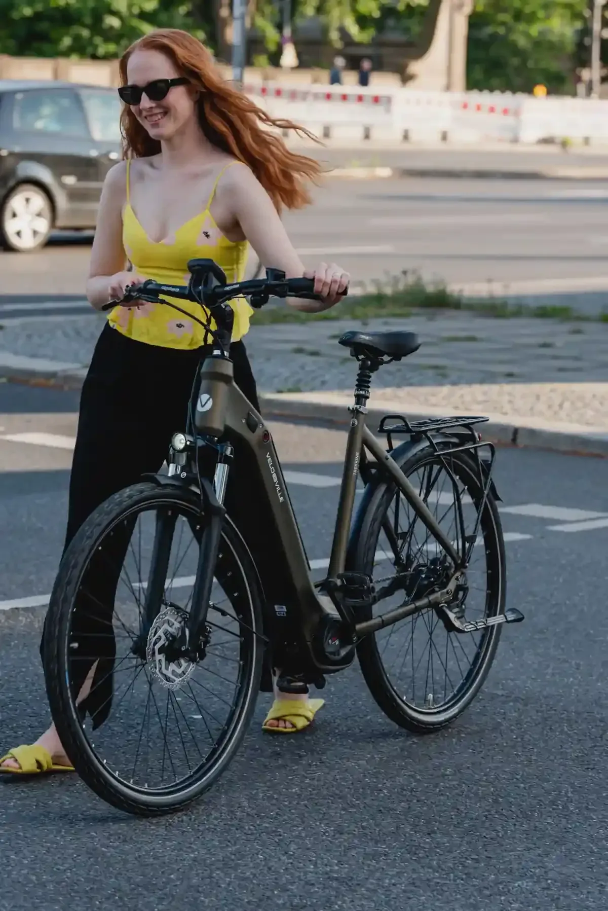 Mujer con blusa amarilla y falda negra de pie junto a una bicicleta en una calle de la ciudad.