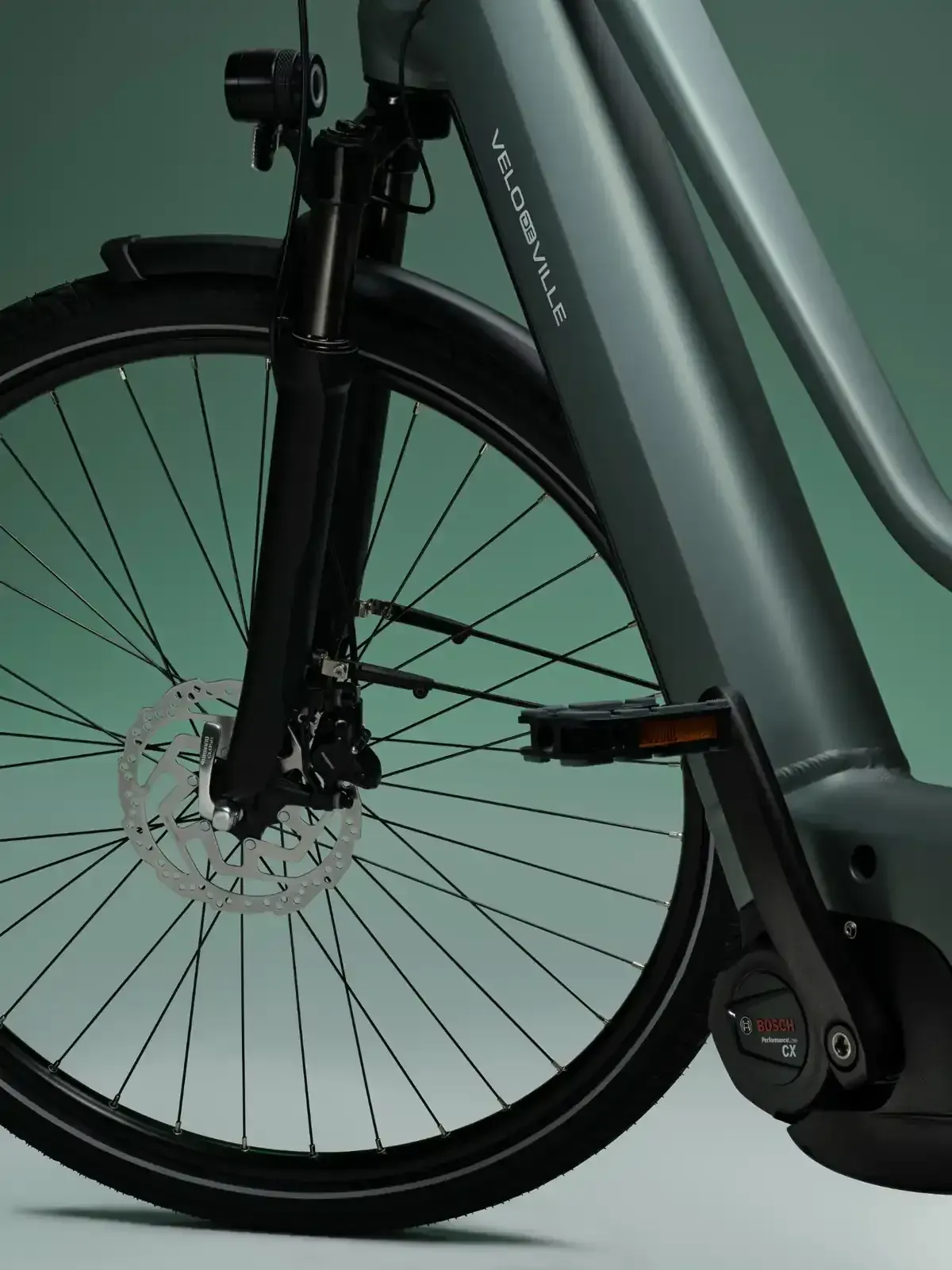 Primer plano de la rueda delantera y la horquilla de una bicicleta con guardabarros y pedal sobre un cuadro gris.