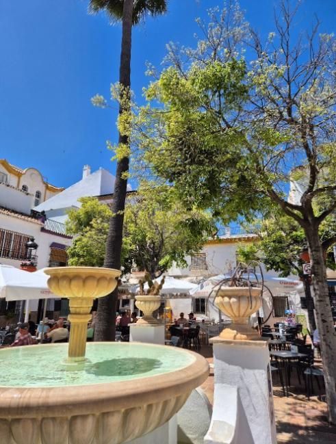 Fuente en una plaza iluminada por el sol con mesas y edificios blancos, cielo azul arriba.