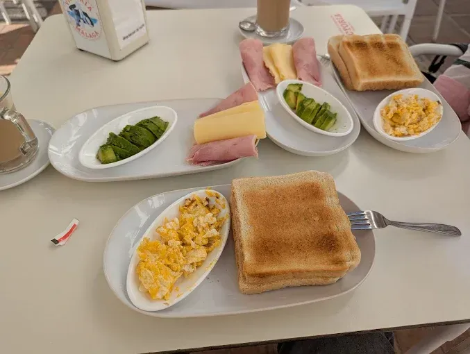 Desayuno: tostadas, huevos revueltos, aguacate, jamón y queso en platos blancos, sobre una mesa blanca.