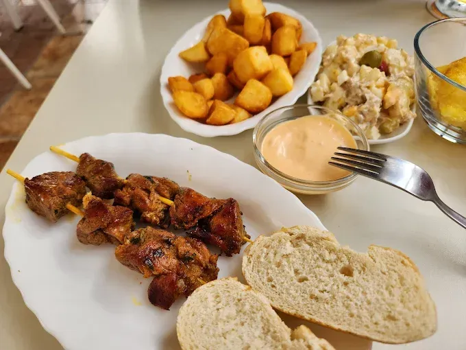 Carne en brocheta, patatas fritas, ensalada de patatas, pan y salsa sobre una mesa blanca.
