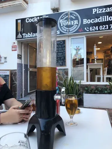 Torre de cerveza y vaso en una mesa al aire libre en un restaurante, persona usando un teléfono, cartel en el fondo.