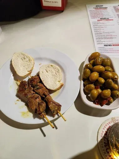 Plato de kebabs con pan, un bol de aceitunas y un menú sobre una mesa blanca.