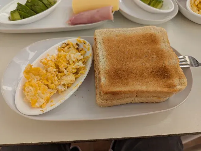 Plato de desayuno con huevos revueltos y tostadas. Otros platos con aguacate, jamón y queso de fondo.