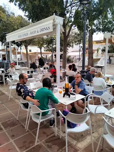 Cafetería al aire libre; gente sentada en mesas, algunos bebiendo. Mesas y sillas blancas, toldo blanco y árboles.