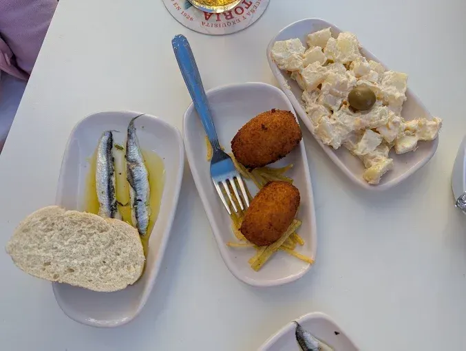 Pequeños platos de tapas sobre una mesa blanca: anchoas, croquetas, ensalada de patatas.