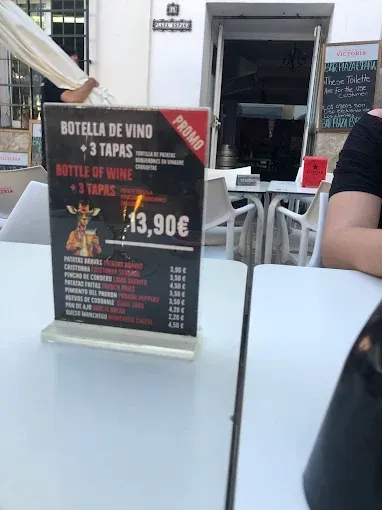 Menú sobre una mesa, anunciando una botella de vino y tres tapas por 13,90 euros. Cafetería al aire libre.