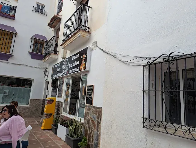 Escena callejera con un edificio blanco, una tienda con cartel y una persona sentada afuera.