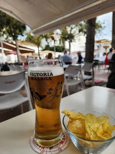 Un vaso de cerveza Victoria Málaga y un bol de patatas fritas sobre una mesa blanca en una cafetería al aire libre.
