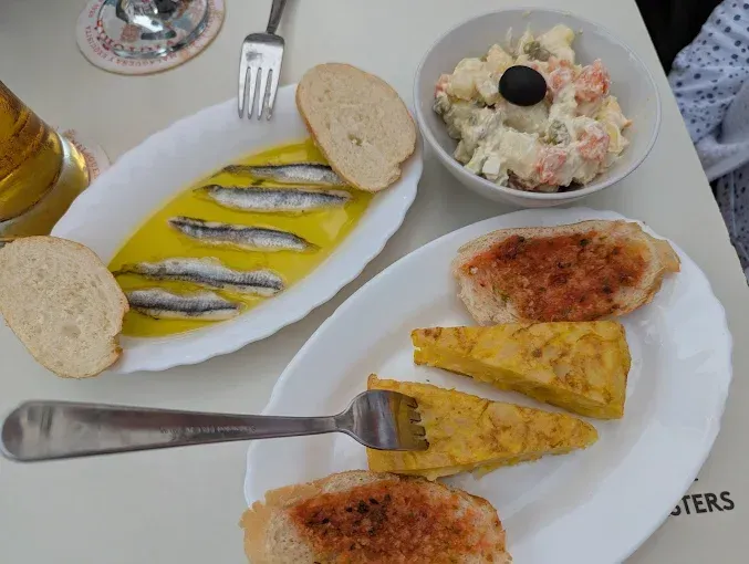 Platos de tapas españolas: anchoas en aceite, ensalada de patatas, tortilla y pan tostado.