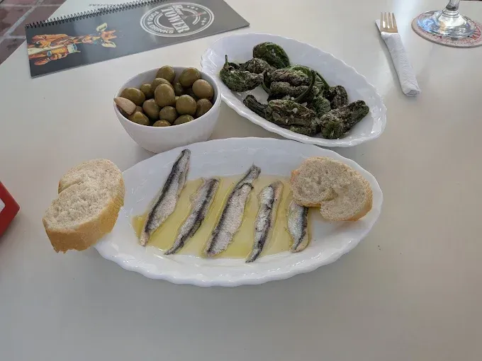 Surtido de tapas: aceitunas, pimientos fritos, anchoas en aceite con pan y menú en mesa.
