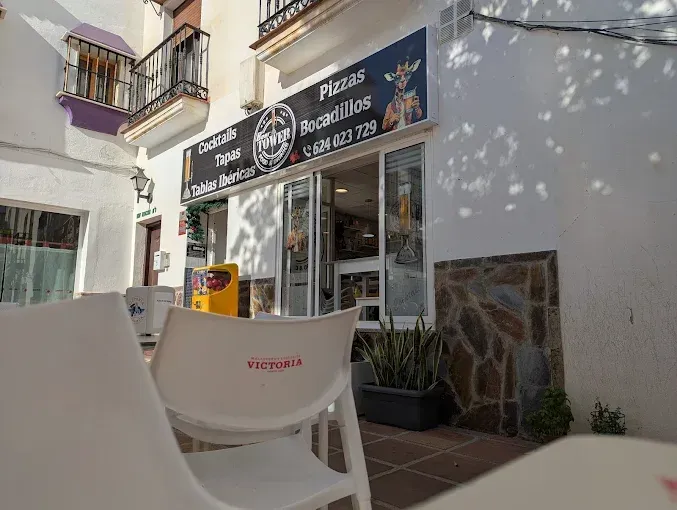 Exterior de un restaurante con sillas blancas y un cartel de pizzas y otros alimentos.