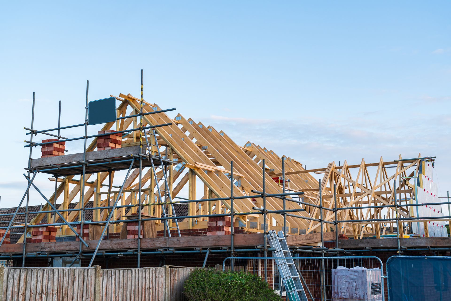 Charpente en bois d'une maison en construction, avec échafaudages et fermes de toit sur fond de ciel bleu.