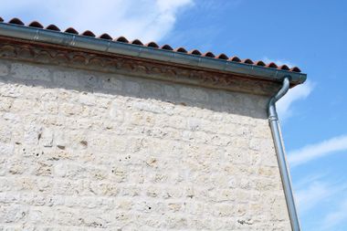 Mur d'un bâtiment en pierre blanche avec un toit de tuiles et une gouttière se détachant sur un ciel bleu