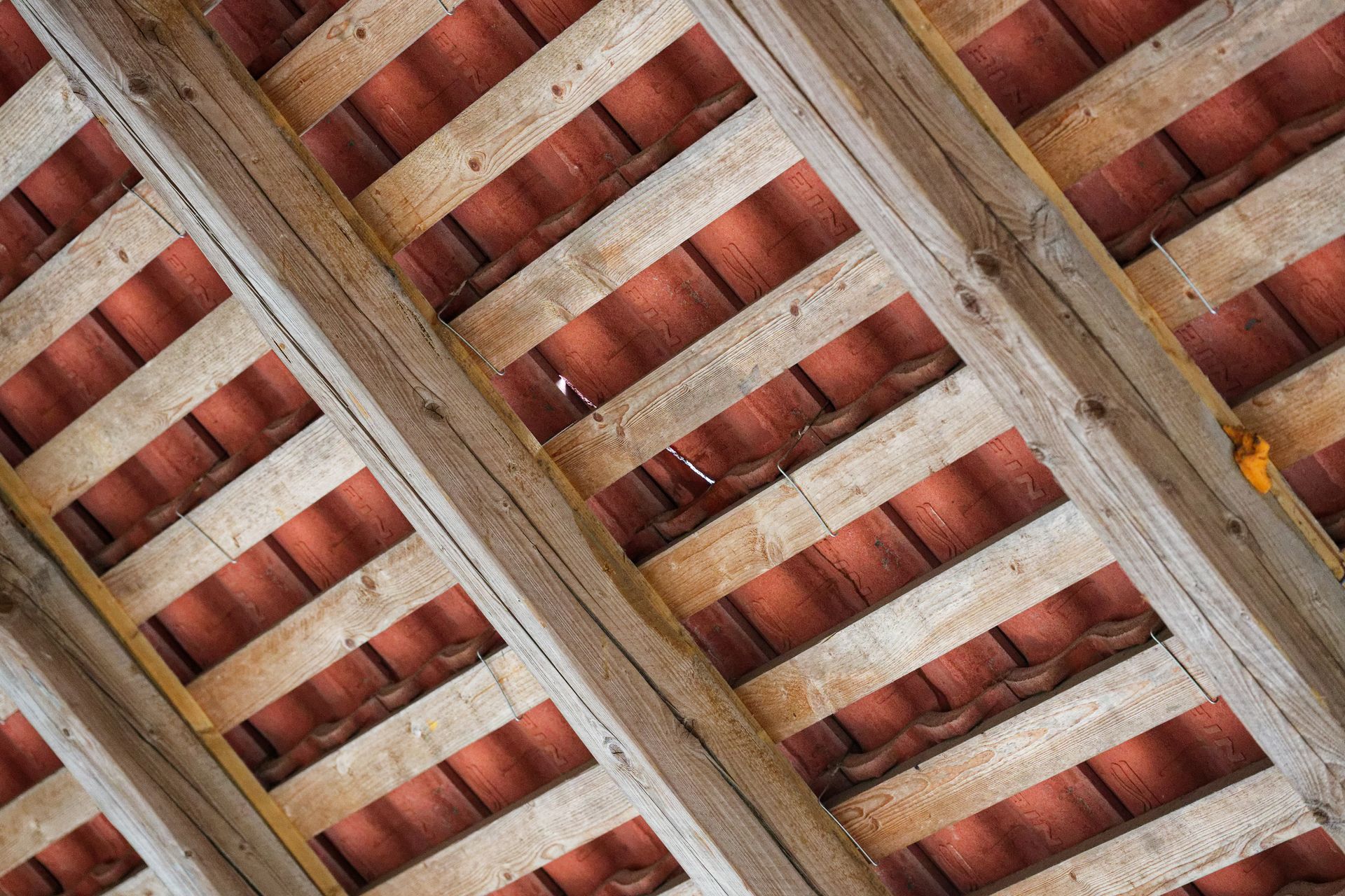 Vue en diagonale de poutres en bois patinées par le temps, avec des tuiles rouges en dessous et quelques feuilles jaunes.