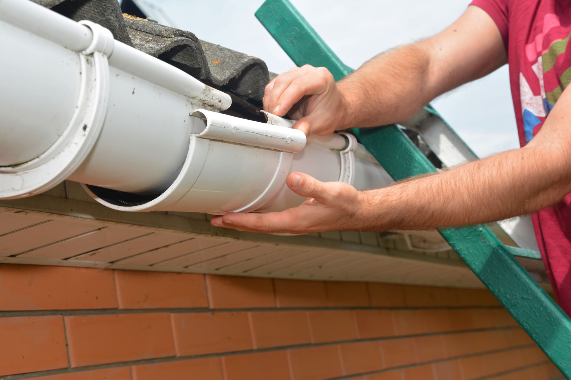 Des mains installent une gouttière blanche sur un toit, à côté d'une échelle verte et d'un mur de briques.