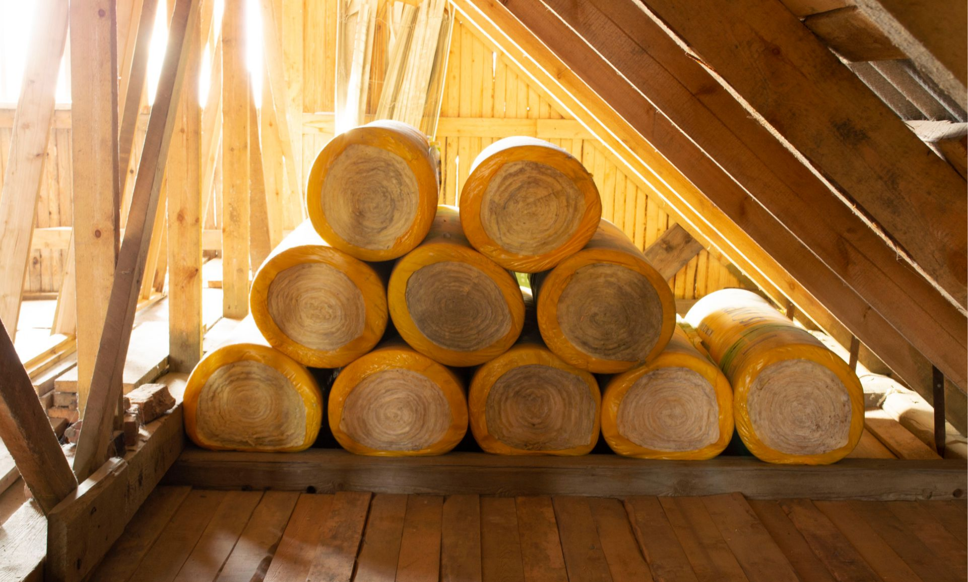 Des rouleaux d'isolant jaune empilés dans un grenier aux poutres en bois apparentes.