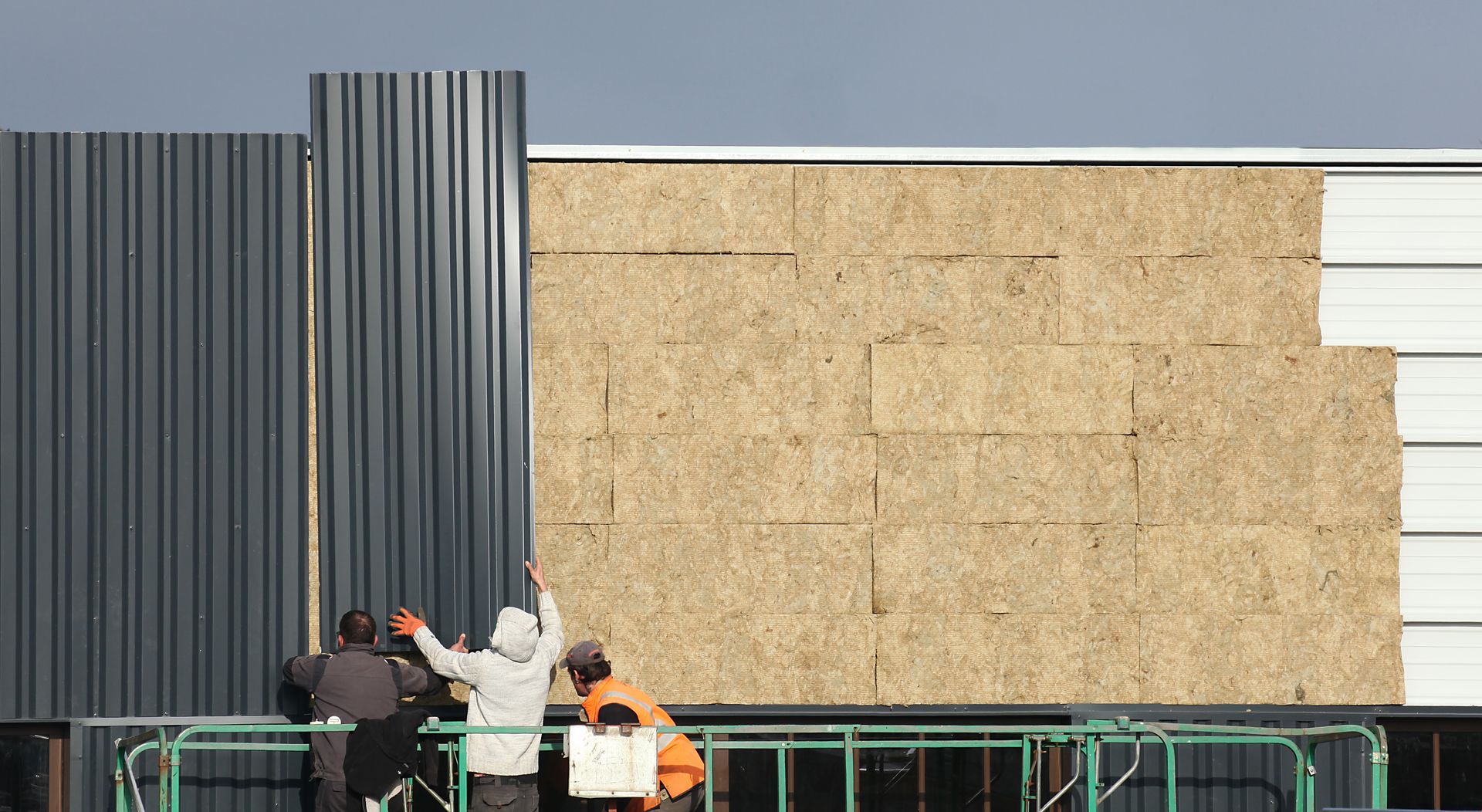 Des ouvriers recouvrent un mur de panneaux beiges à côté d'un bâtiment à rayures grises et de barricades vertes.