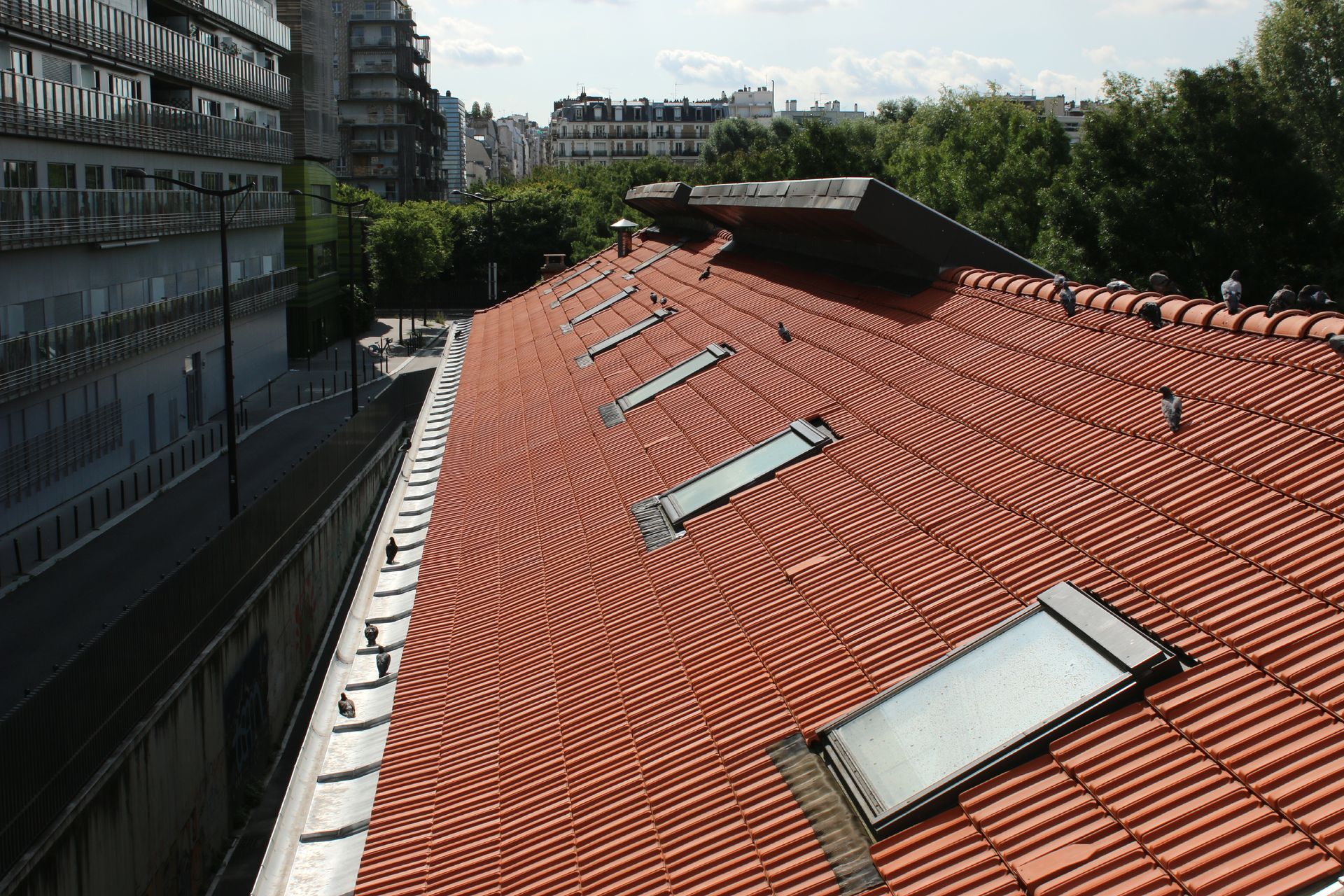 Toit de tuiles rouges avec puits de lumière à côté d'un bâtiment moderne et d'arbres sous un ciel partiellement nuageux