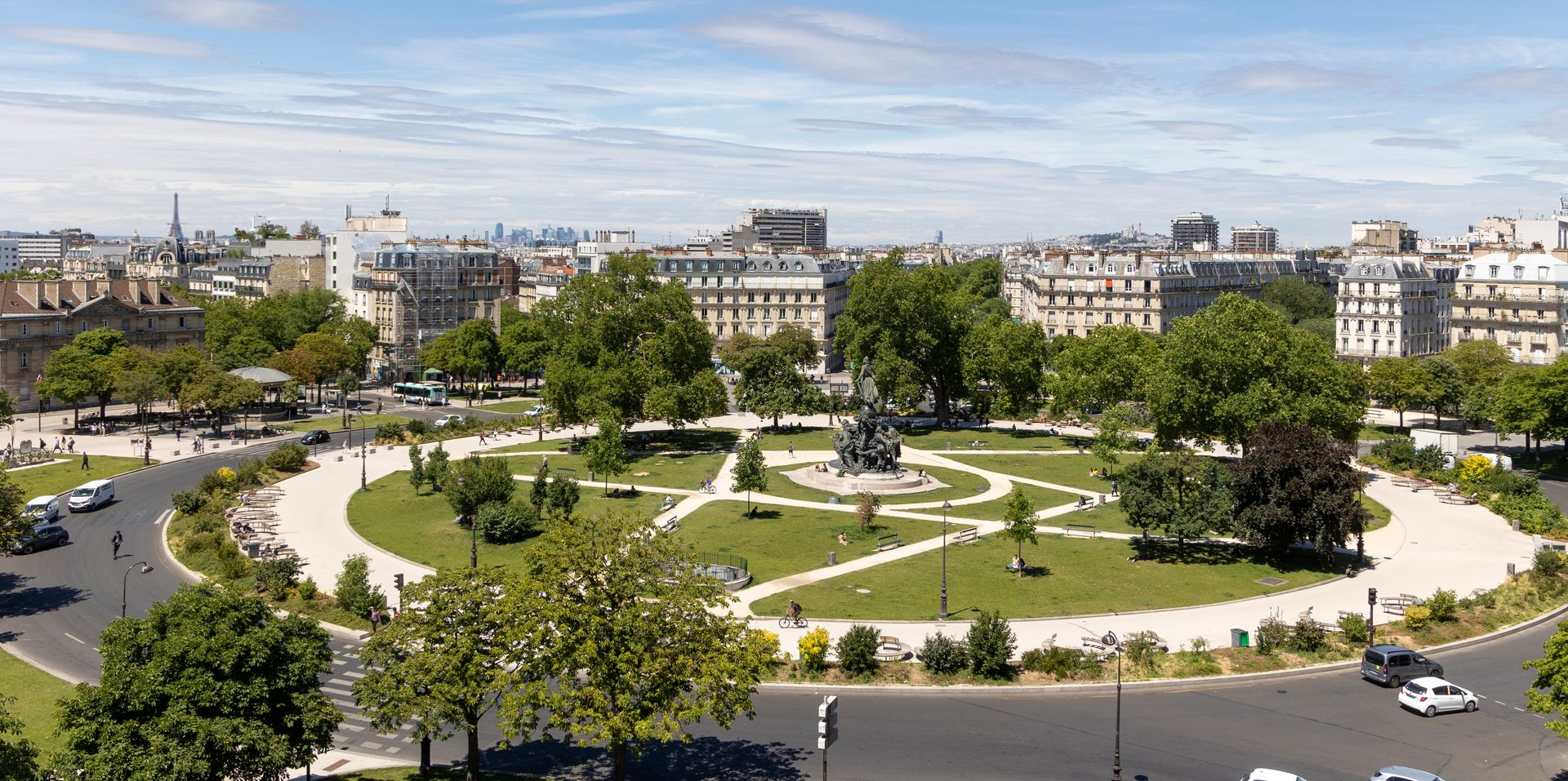 Vue aérienne d'un parc urbain verdoyant avec des sentiers sinueux, des arbres et des bâtiments environnants.