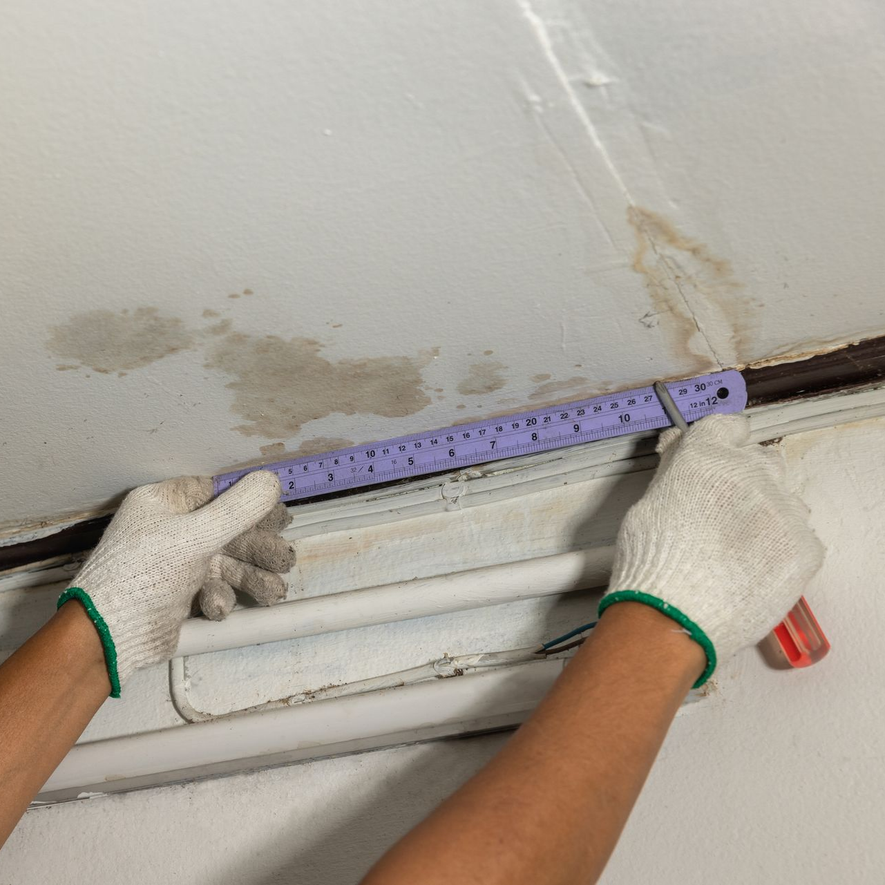 Des mains gantées mesurent un mur abîmé avec une règle violette près d'une peinture écaillée et d'une fissure.