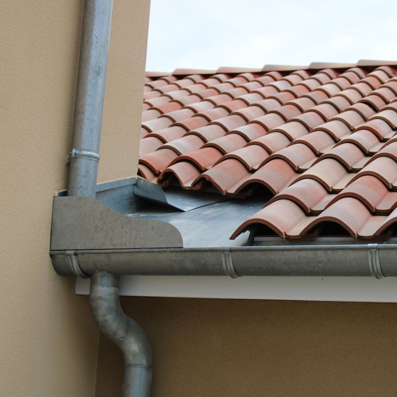 Gouttière et descente pluviale sur une maison beige avec des tuiles en terre cuite rouge et un solin de noue