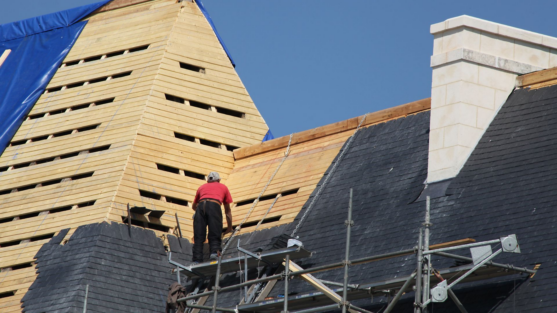 Des ouvriers sur des échafaudages posent un toit de bardeaux de bois sur une maison, sous une bâche bleue.