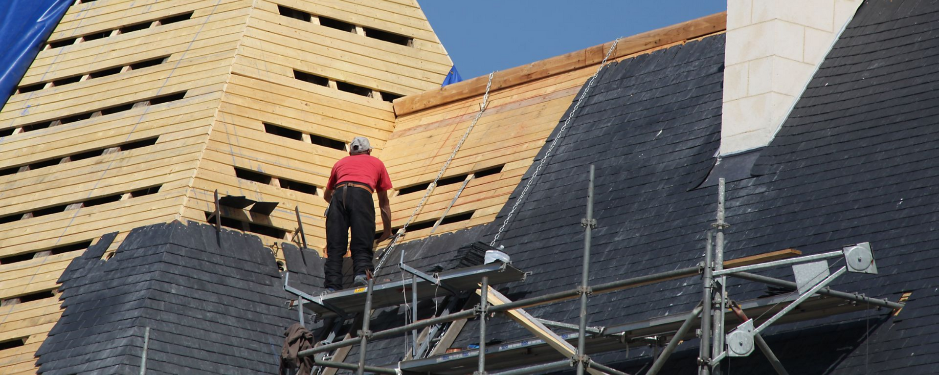 Un ouvrier en veste rouge se tient sur un échafaudage à côté d'un bâtiment en construction au toit pentu.
