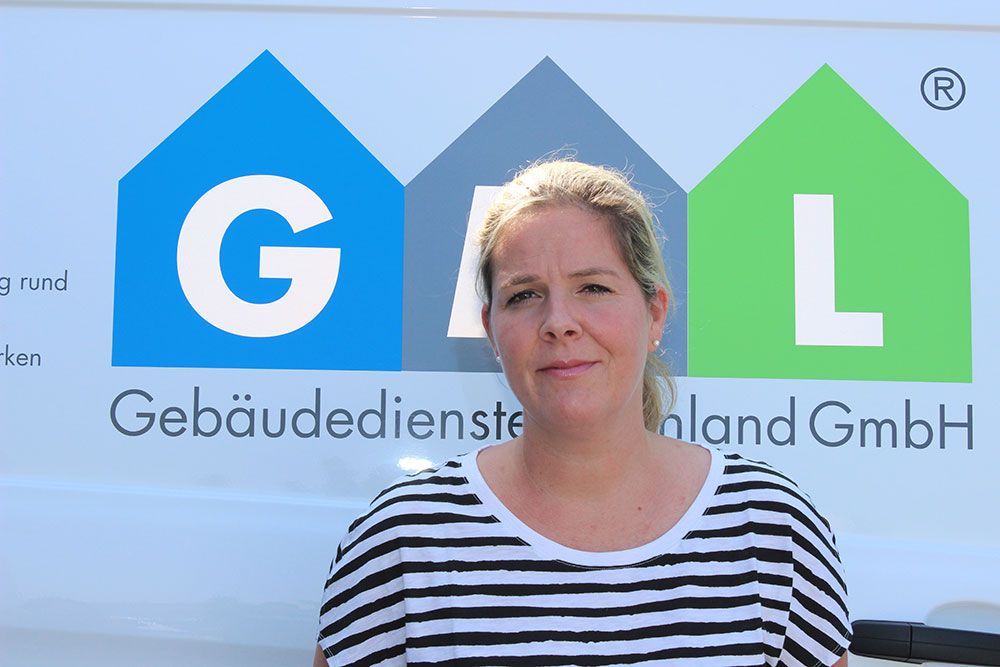 Eine Frau in einem gestreiften Hemd steht vor einem Schild mit der Aufschrift „Gebäudedienste Land GmbH“.