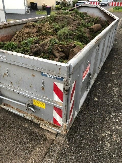 Am Straßenrand steht ein mit Erde und Gras gefüllter Müllcontainer.