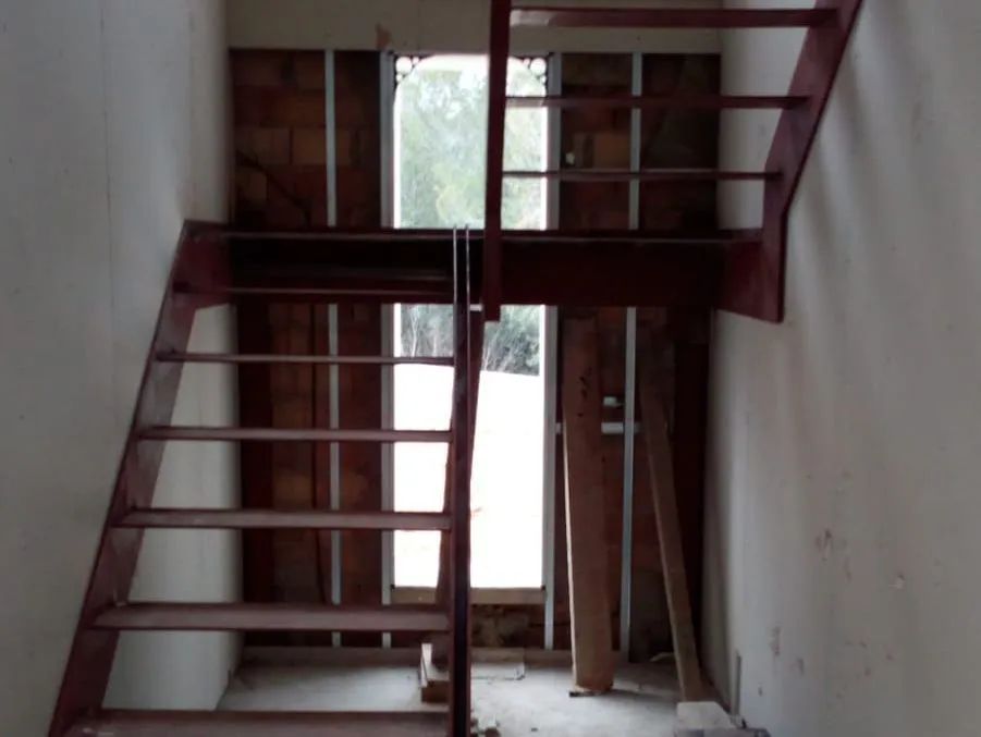 Escalera de madera en un espacio interior sin terminar, con paredes blancas y vigas a la vista.