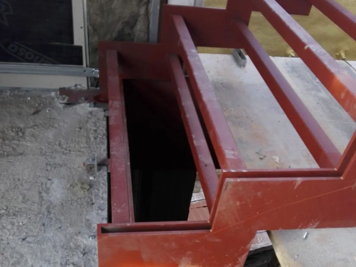 Abertura de escalera metálica roja junto a un suelo de hormigón, con escalones de madera encima.