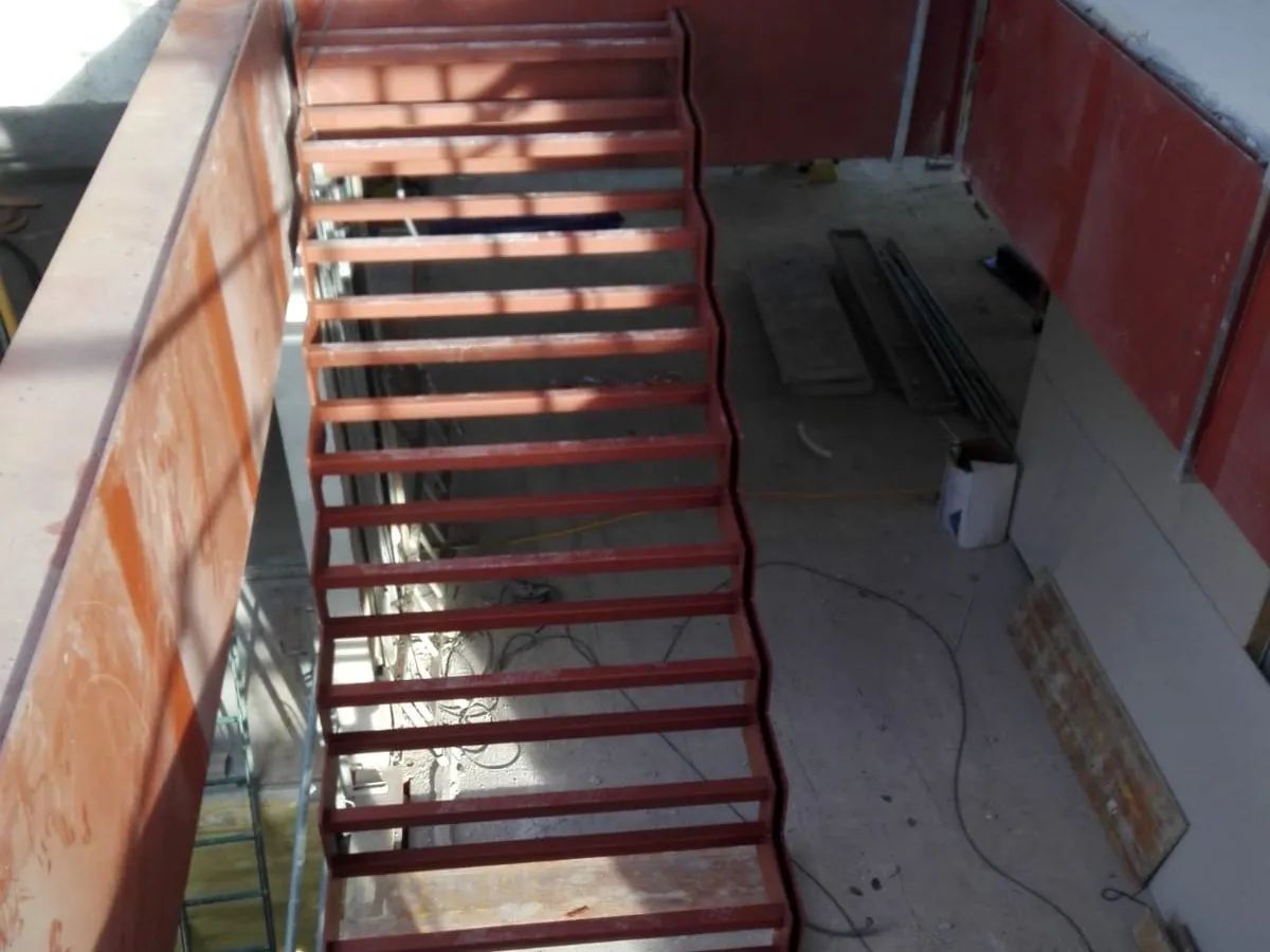 Estrecha escalera exterior con escalones y barandillas de metal rojo junto a una pared roja.