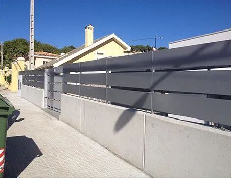 Acera residencial junto a una pared blanca y una valla metálica gris bajo un cielo azul claro.