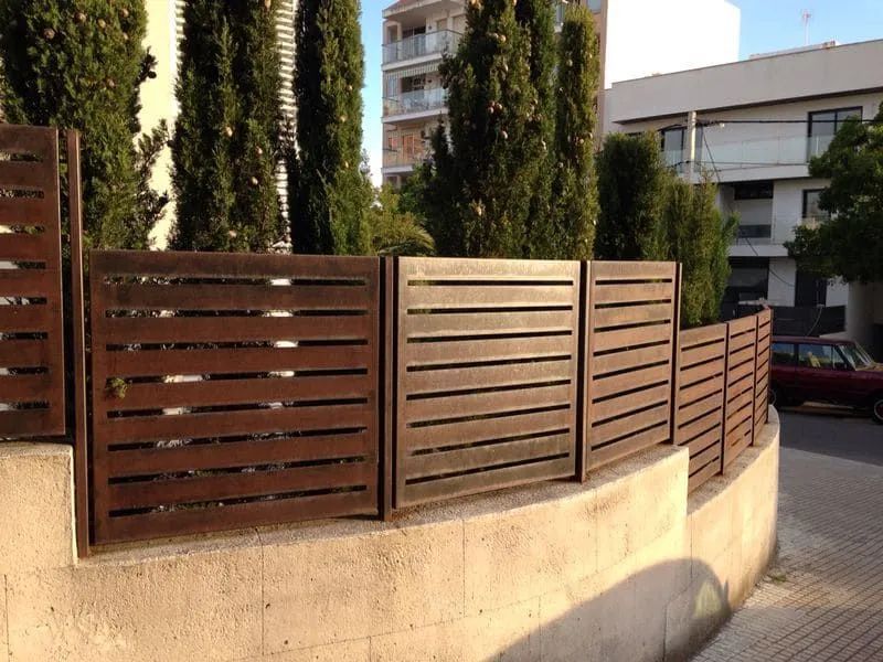 Una valla curva de madera se alza sobre un muro de hormigón junto a una calle