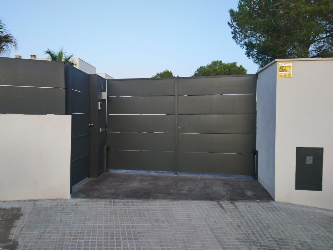 Puerta de entrada moderna de color gris con paredes blancas y primer plano pavimentado bajo un cielo azul.