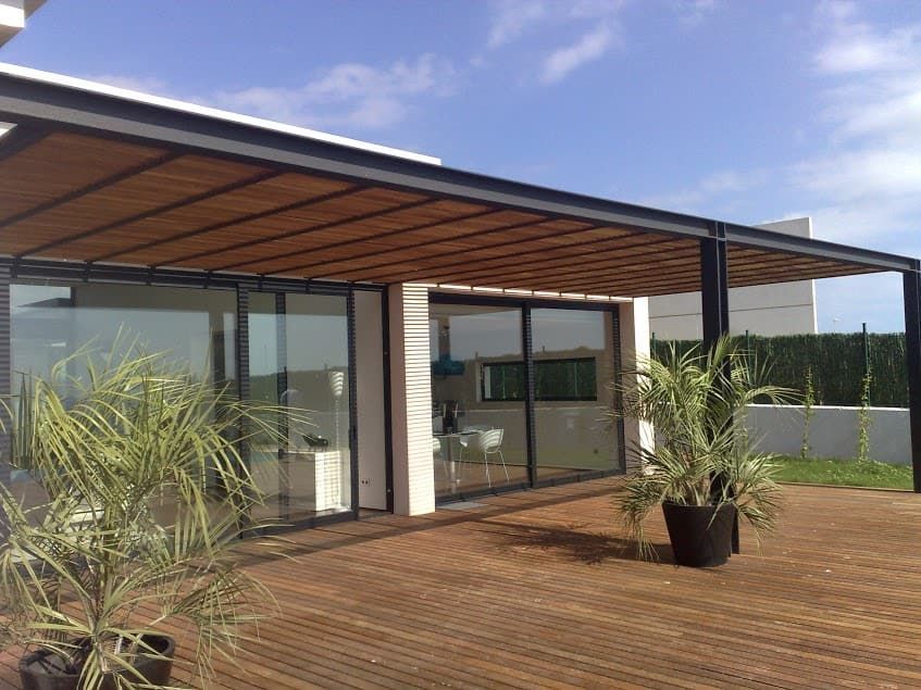 Patio de una casa moderna con terraza de madera, paredes de cristal, plantas en macetas y techo de listones