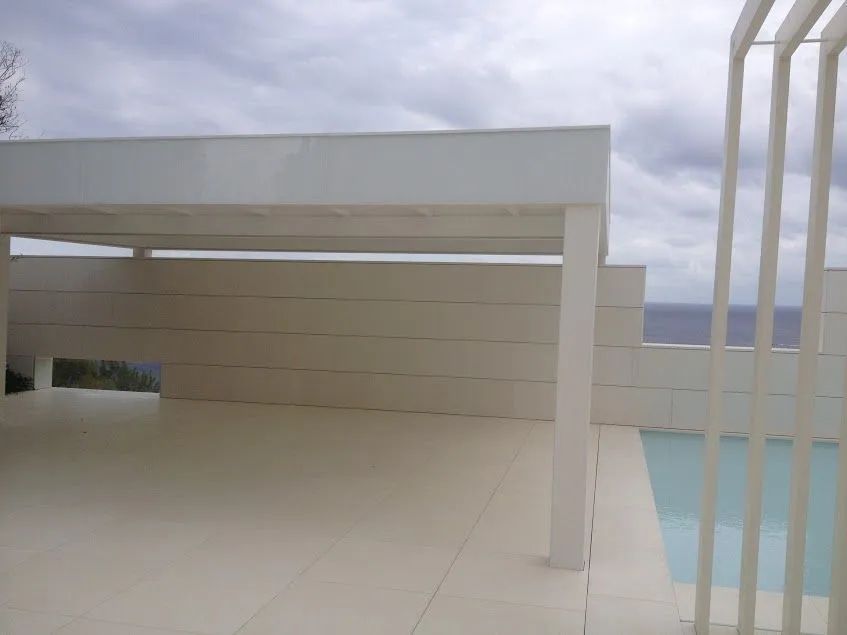 Terraza blanca en la azotea con pérgola, suelo de baldosas y barandilla de piscina bajo un cielo nublado.