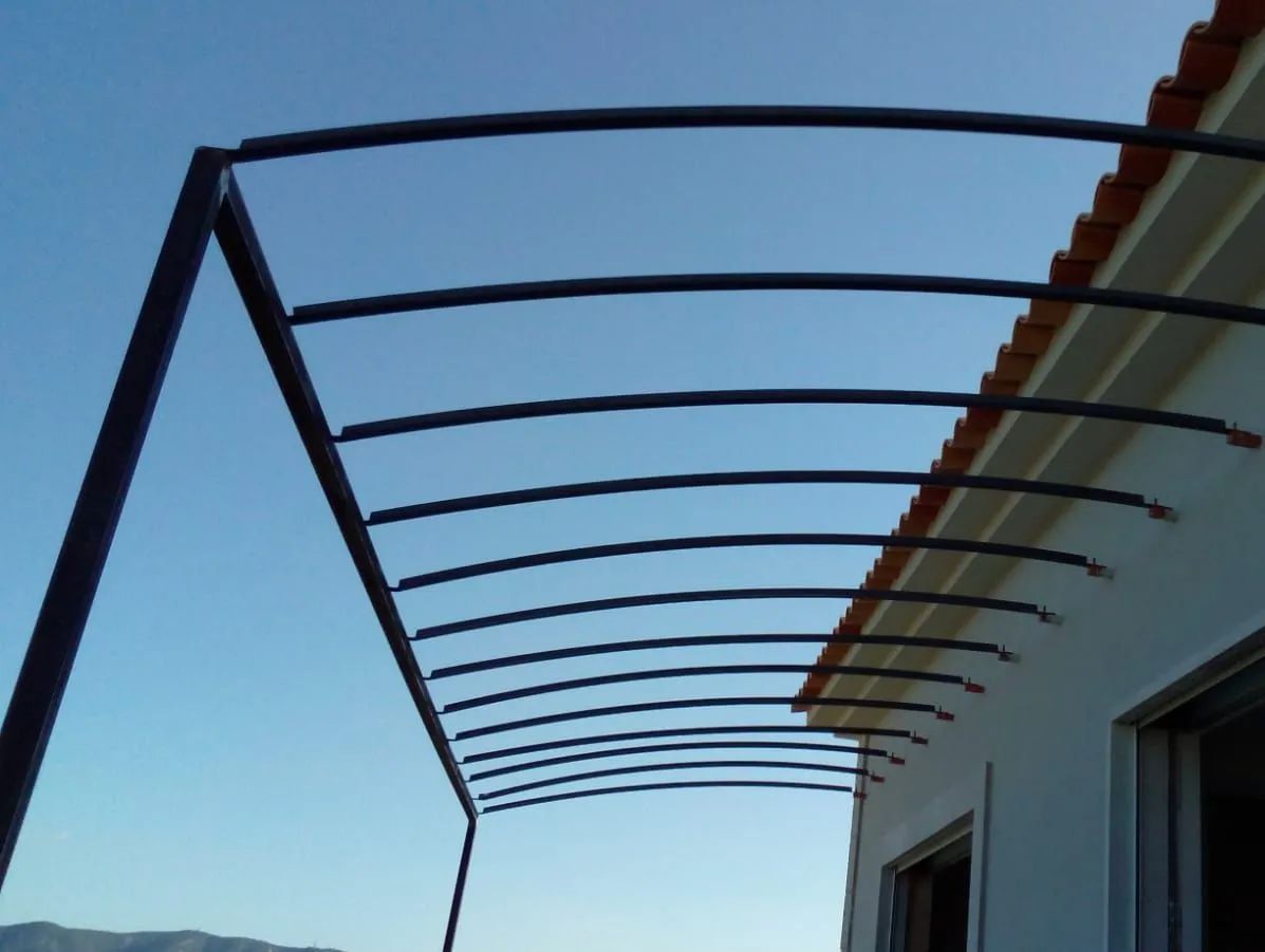 Estructura metálica de un toldo que se extiende desde un edificio blanco bajo un cielo azul despejado.