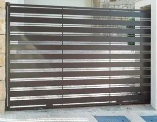 Puerta de metal oscura con listones horizontales frente a la entrada de un edificio, con acera y puertas de vidrio detrás.