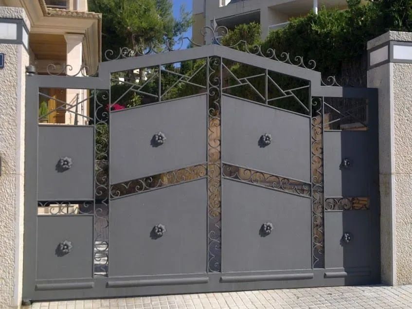 Puerta de entrada moderna de metal gris con paneles geométricos decorativos y pilares de piedra.