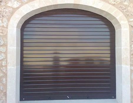 Persiana metálica oscura cerrada, colocada en un arco de piedra con una vista borrosa detrás.