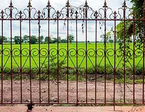 Valla metálica ornamentada con un campo verde y árboles al fondo.