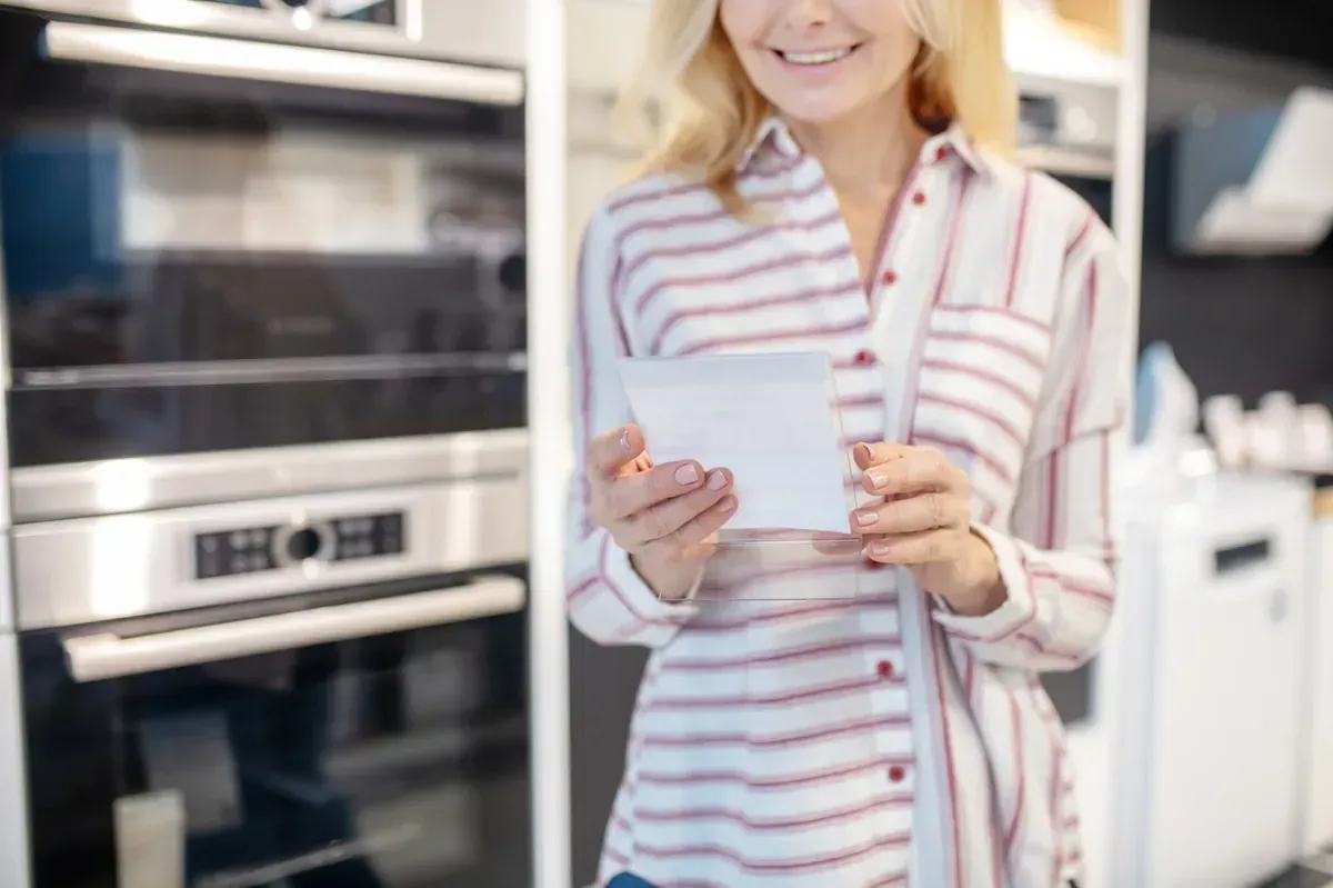 Mujer con camisa de rayas sonriendo, sosteniendo papel, en una tienda de electrodomésticos de cocina, mirando hornos.