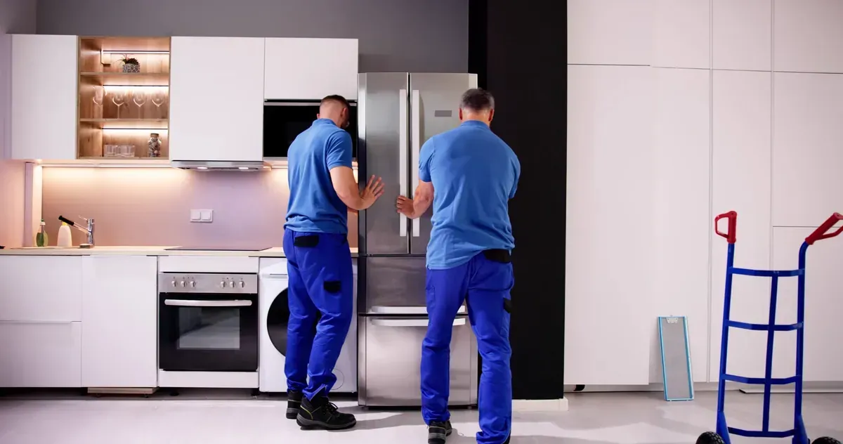Dos operarios con uniformes azules transportan un frigorífico de acero inoxidable en una cocina moderna.