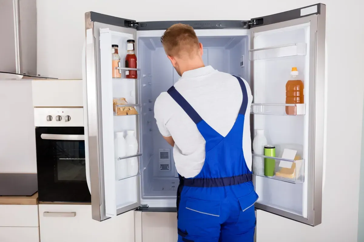 Reparador con mono azul inspeccionando el interior de un refrigerador con las puertas abiertas.