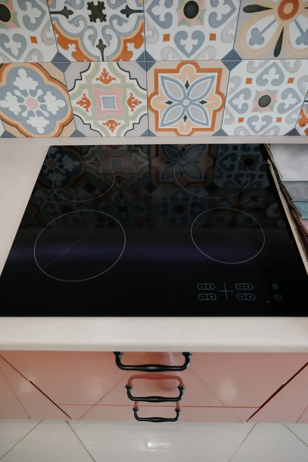 Cocina eléctrica negra con cuatro quemadores, ubicada sobre una encimera blanca frente a un protector contra salpicaduras de azulejos estampados y cajones.
