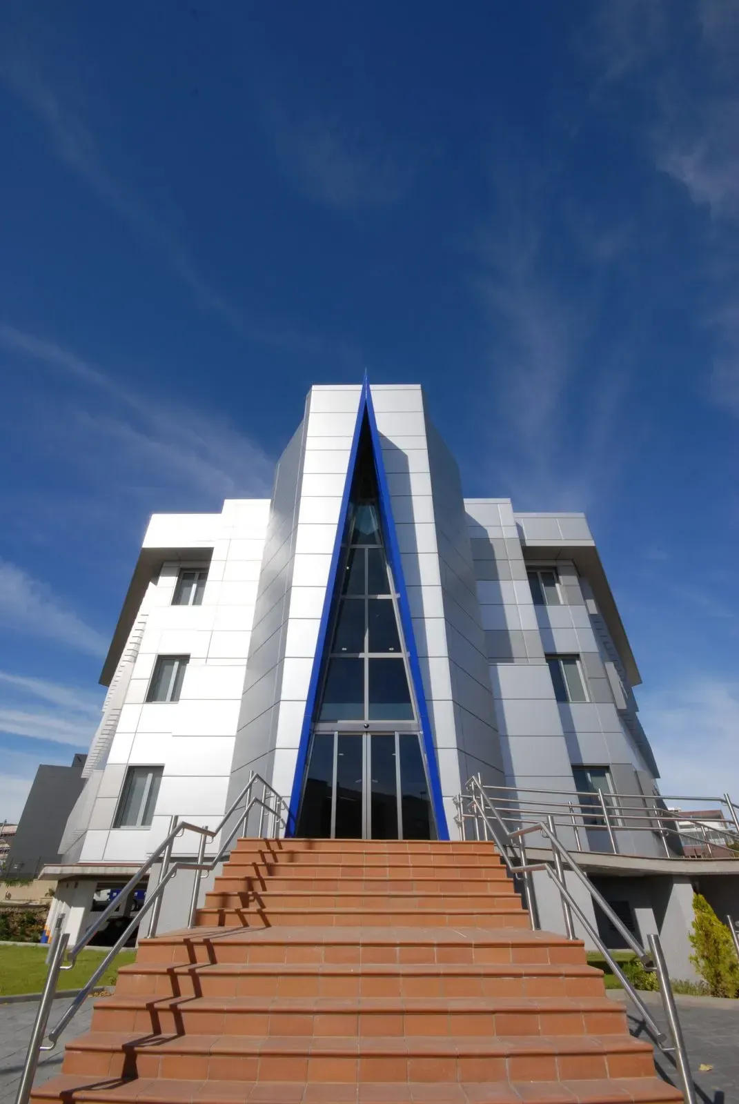Edificio moderno revestido de plata con una entrada triangular de cristal, situado en el centro sobre una amplia escalera naranja bajo un cielo azul.